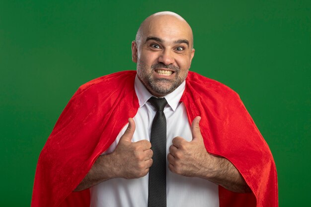 Super hero businessman in red cape looking at front happy and positive smiling showing thumbs up standing over green wall