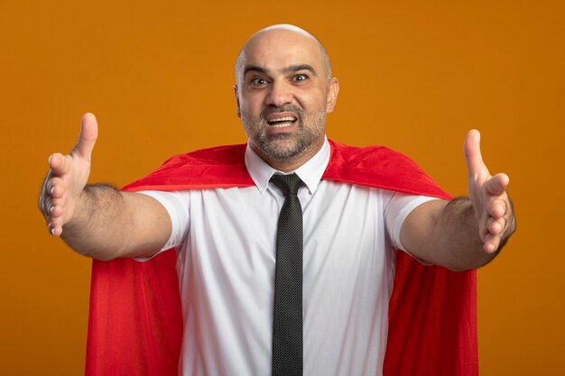 Super hero businessman in red cape looking at front smiling friendly making welcoming gesture with hands standing over orange wall