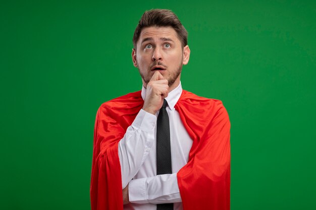 Super hero businessman in red cape looking up puzzled standing over green wall