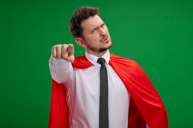 Super hero businessman in red cape pointing with index figner at camera being displeased standing over green background