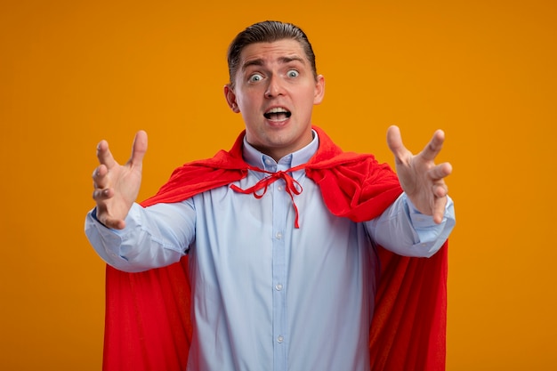 Free photo super hero businessman in red cape  with crazy look of surprisebeing crazy amazed and surprised with arms out standing over orange wall