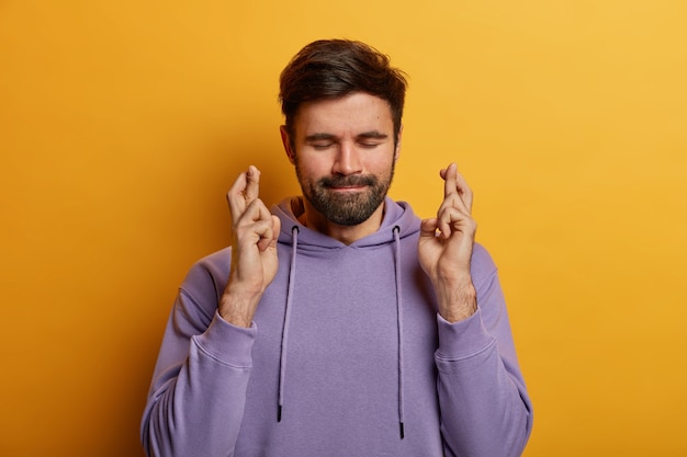 Free photo superstitious bearded young man crosses fingers for good luck, hopes wish comes true, closes eyes and presses lips, has big desire or dream, wears purple sweatshirt, isolated on yellow wall