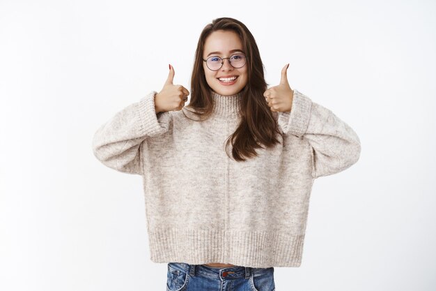 Supportive and optimstic glad nice european female brunette in glasses and sweater showing thumbs up impressed and delighted, liking product giving positive answer, recommending it over gray wall.