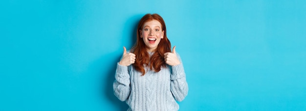 Supportive redhead girl in sweater showing thumbsup and looking amazed praising good choice standing