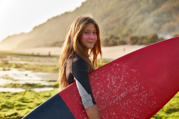 Surfer and ocean concept. Delighted dark haired woman carries waxed surfboard looks with satisfied expression