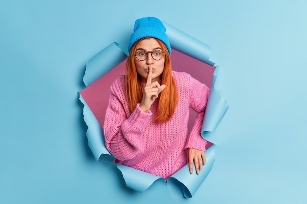 Free photo surpised redhead young woman looks mysteriosly keeps index finger over lips tells secret information gossips about something wears hat and pink sweater.