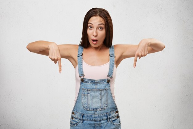 Surprise, shock and astonishment concept. Shocked pretty woman in denim jumpsuit, pointing both index fingers down, indicating something astonishing