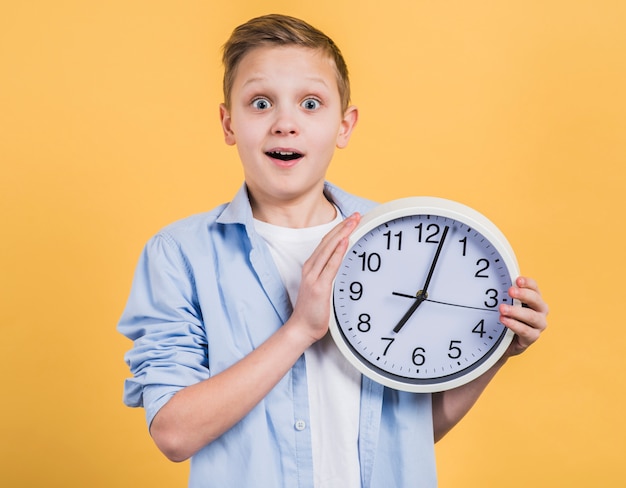 Surprise smiling boy holding white clock in hand looking to camera against yellow background