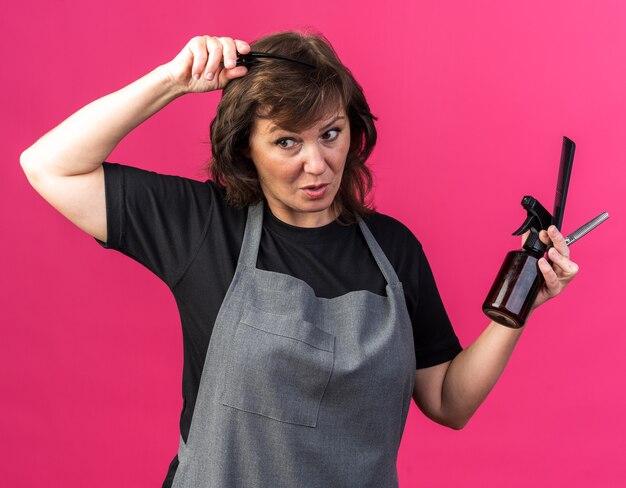 Surprised adult caucasian female barber in uniform holding barber tools isolated on pink background with copy space