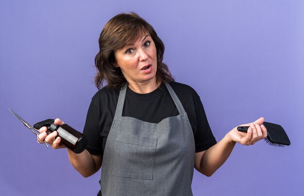 Surprised adult caucasian female barber in uniform holding spray bottle comb and scissors isolated on purple wall with copy space