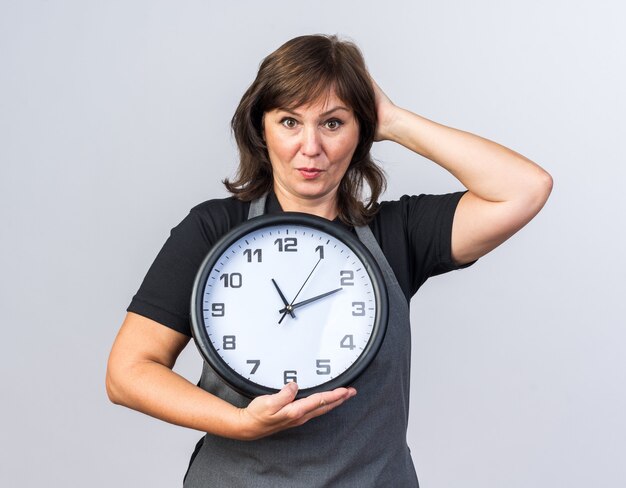 surprised adult female barber in uniform putting hand on head and holding clock isolated on white wall with copy space