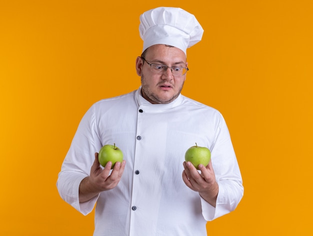 Free photo surprised adult male cook wearing chef uniform and glasses holding apples looking at one of them isolated on orange background