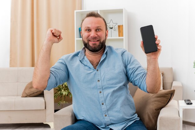 Surprised adult slavic man sits on armchair keeping fist up holding phone inside the living room