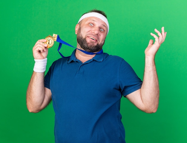 surprised adult slavic sporty man wearing headband and wristbands holding golden medal isolated on green wall with copy space