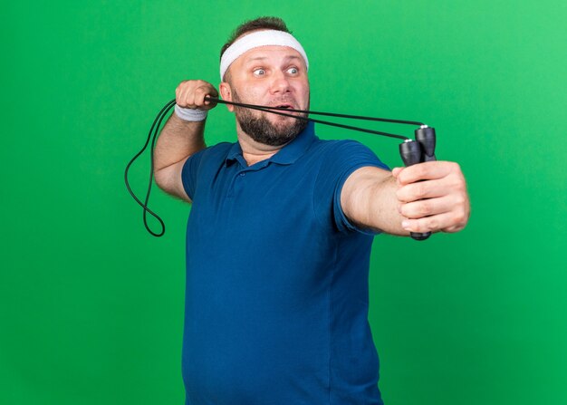 surprised adult slavic sporty man wearing headband and wristbands holding jumping rope and looking at side isolated on green wall with copy space