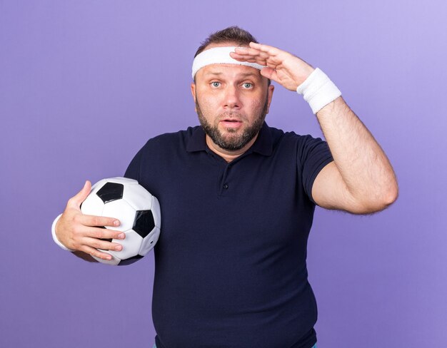 surprised adult slavic sporty man wearing headband and wristbands keeping palm at forehead and holding ball isolated on purple wall with copy space