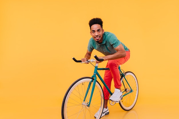 Free Photo surprised african guy in red pants riding on bike. indoor photo of funny black young man sitting on bicycle.