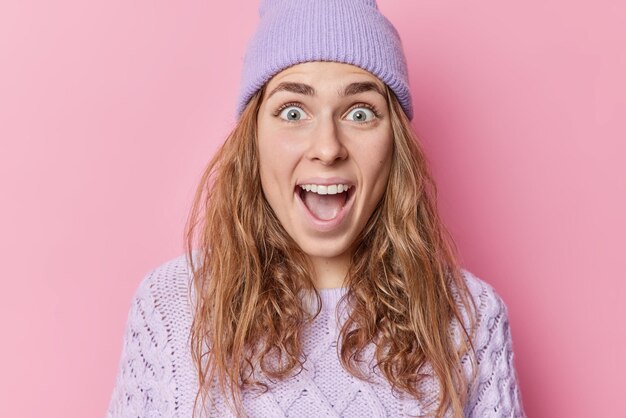 Surprised amazed European woman keeps mouth opened stares bugged eyes reacts on unexpected surprise drops jaw wears hat and sweater isolated over pink background. People and reactions concept