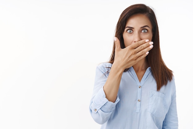 Surprised amused middle-aged woman brunette shocked stare at front, cover mouth speechless, press palm lips astonished, gossiping, hear stunning rumor, pose white wall