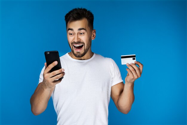 Surprised attractive guy is watching on the cell phone and holding credit card