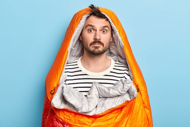 Free Photo surprised bearded male camper poses in sleeping bag, wears striped jumper, spends weekend actively, poses over blue wall, has expedition near rocky mountains