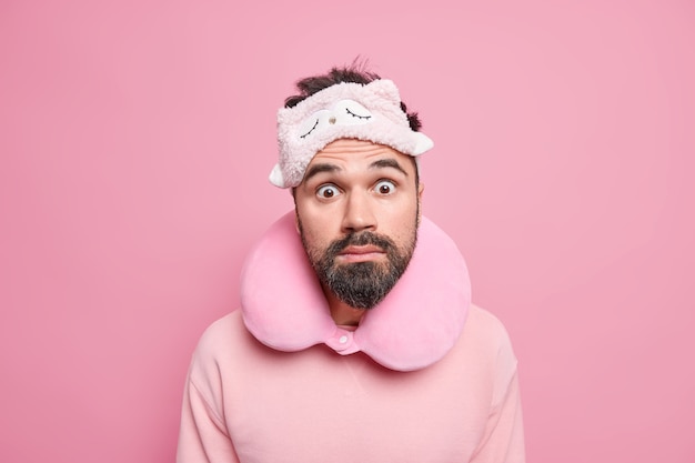 Surprised bearded male tourist wears neckpillow sleepmask casual jumper stares shocked