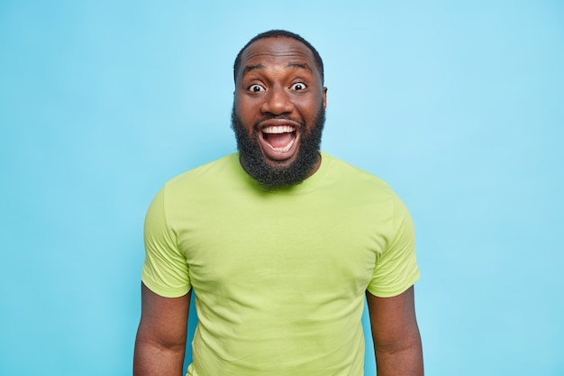 Surprised bearded man reacts on something unexpected keeps mouth opened dressed in casual green t shirt hears excellent news isolated over blue wall