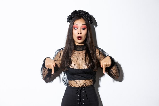 Surprised beatiful asian woman in black gothic dress and wreath, pointing fingers down and looking startled at something