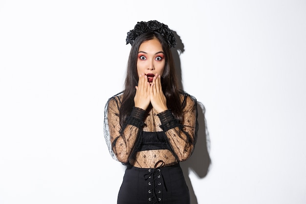 Free photo surprised beautiful asian woman in witch costume looking amazed. female wearing halloween gothic dress and wreath, open mouth aamused, standing over white background.