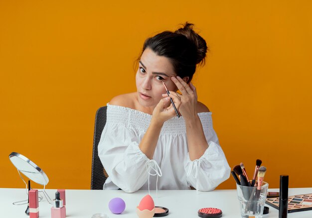 Surprised beautiful girl sits at table with makeup tools applying eyeshadow with makeup brush looking isolated on orange wall