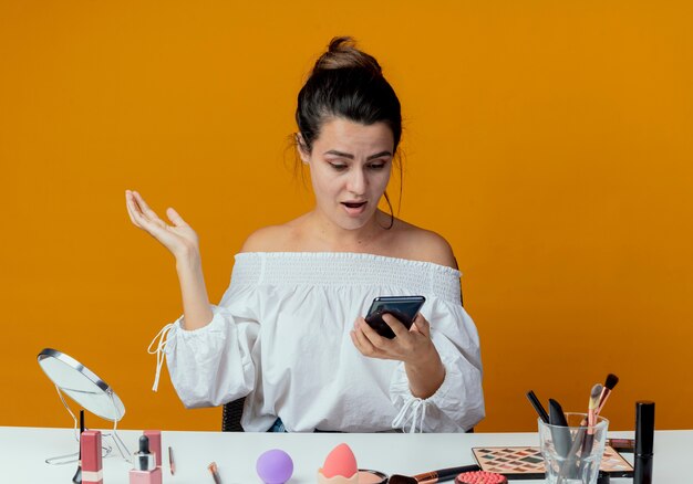 Surprised beautiful girl sits at table with makeup tools holds and looks at phone isolated on orange wall