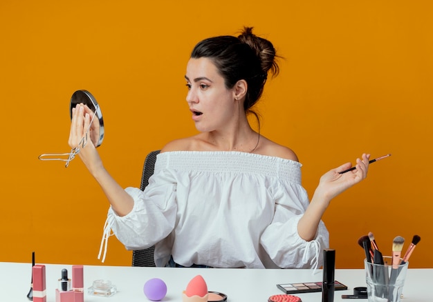 Surprised beautiful girl sits at table with makeup tools looks at mirror and holds makeup brush isolated on orange wall