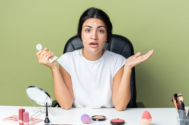 Surprised beautiful woman sits at table with makeup tools applying tone cream spreading hands