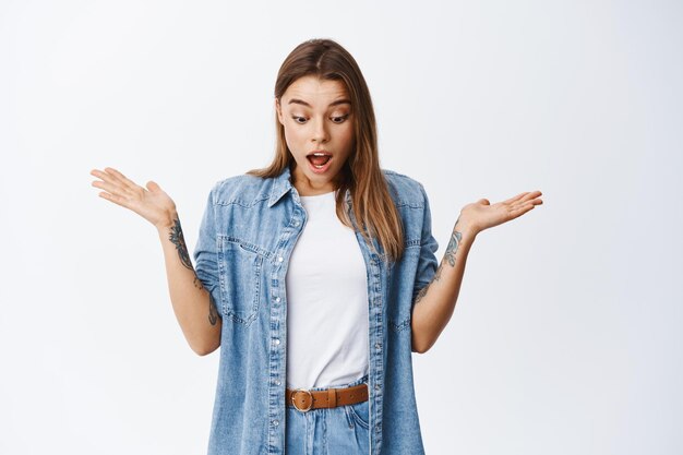 Surprised blond girl gasping in disbelief, raising hands sideways and looking down with shocked face, say wow, impressed by bottom advertisement