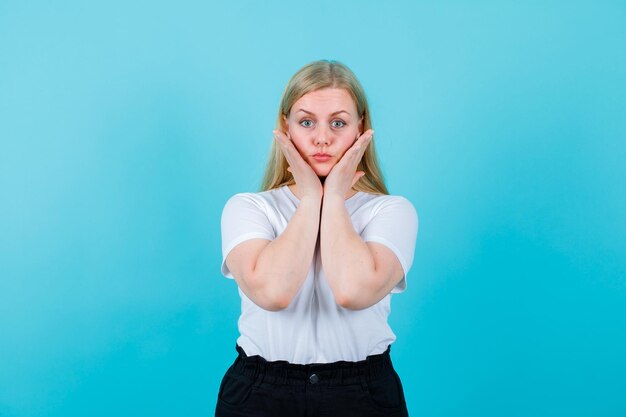 Surprised blonde girl is looking at camera by putting hands on cheeks on blue background