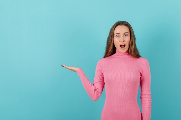 Surprised blonde girl is pointing to left with hand on blue background