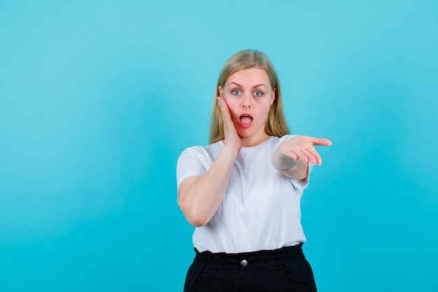 Free Photo surprised blonde girl is putting one hand on cheek and extending other to camera on blue background