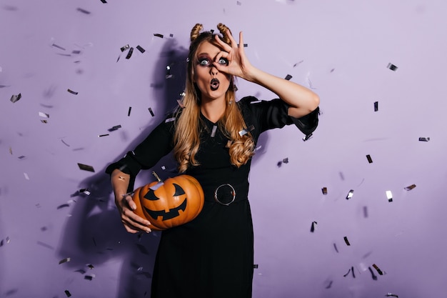 Surprised blonde witch standing under confetti. Shocked young woman posing on purple wall with halloween pumpkin.