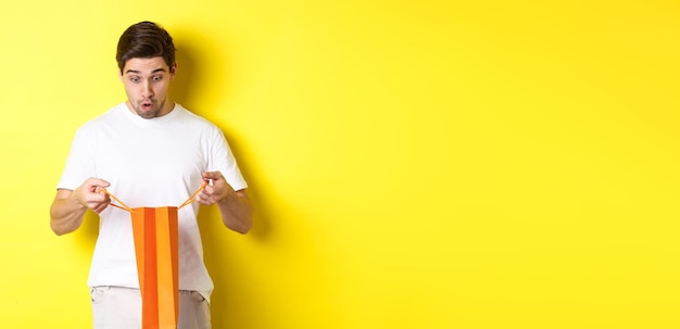 Free Photo surprised boyfriend open bag with gift looking inside amazed standing over yellow background