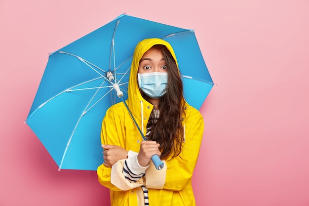 Surprised brunette Asian woman wears medical mask, being protected from catching disease, wears yellow raincoat, holds umbrella