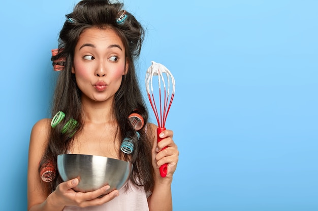 Free photo surprised brunette woman whisks sour cream in bowl, wears hair curlers