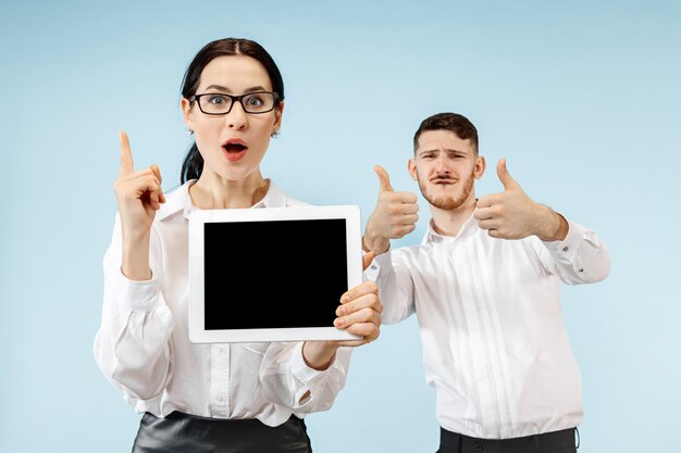 The surprised business man and woman smiling on a blue studio background and showing empty screen of laptop or tablet