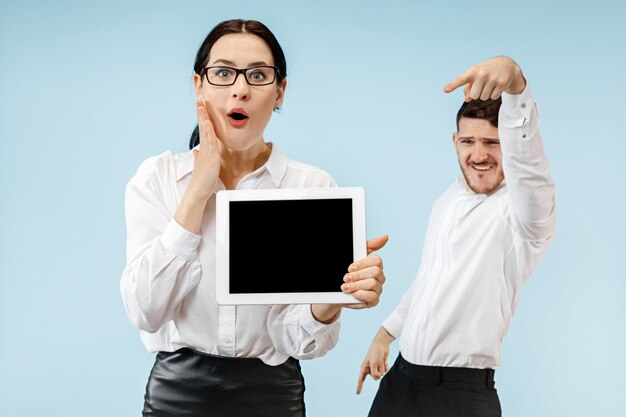 The surprised business man and woman smiling on a blue  wall and showing empty screen of laptop or tablet