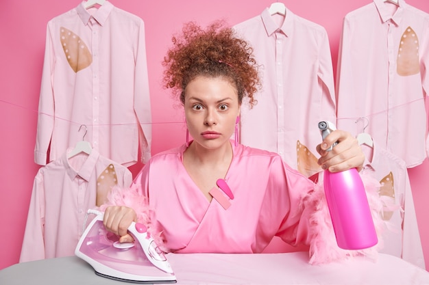 Free photo surprised busy curly haired european housewife holds detergent spray and electric iron uses electric appliance dressed in domestic gown isolated over pink wall with hanging ironed shirts