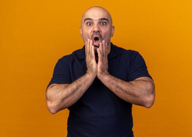 Surprised casual middle-aged man keeping hands on chin looking at front isolated on orange wall