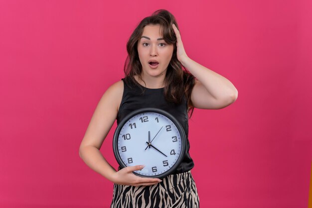 Surprised caucasian girl wearing black undershirt holding a wall clock and put her hand on head on pink background