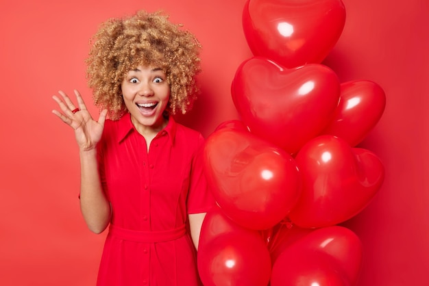 Surprised cheerful curly haired woman gets uexpected surprise gify on Valentines Day holds bunch of heart shaped balloons wears dress isolated over red background People and celebration concept