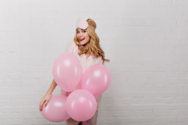 Free photo surprised cheerful girl in sleepmask standing on white wall with party balloons. portrait of beautiful young lady celebrating something in weekend morning.