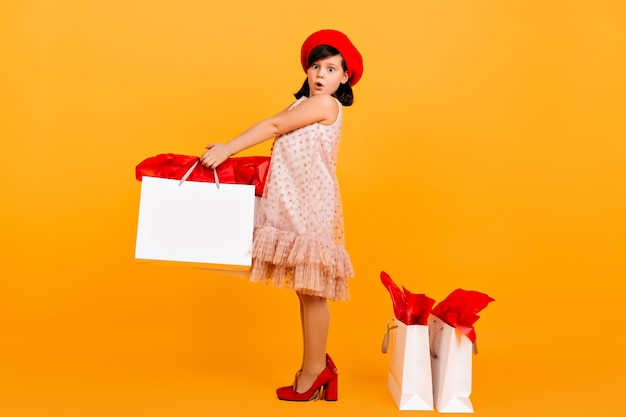 Surprised child posing in mother's shoes.  amazed preteen girl holding shopping bag on yellow wall.