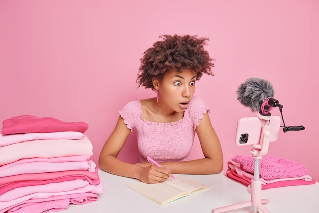 Surprised curly haired Afro American woman makes notes in notebook focused at smartpphone camera sits at table surrounded by piles of neatly folded laundry makes video blog about domestic routine